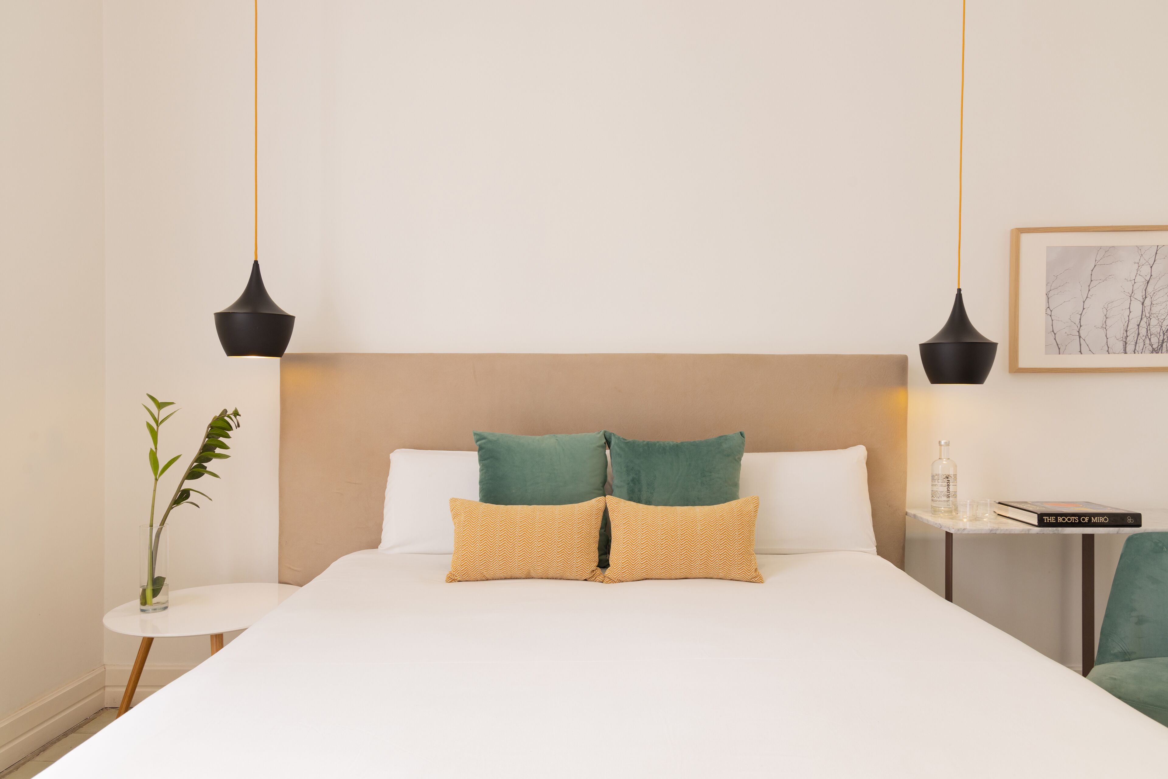 Habitación de hotel ordenada con una cama, dos mesitas de noche, una mesita auxiliar pequeña, una planta en maceta y dos lámparas colgantes.