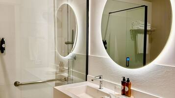 A modern bathroom with a glass shower enclosure, a large mirror with a circular frame, and a white sink with toiletries.