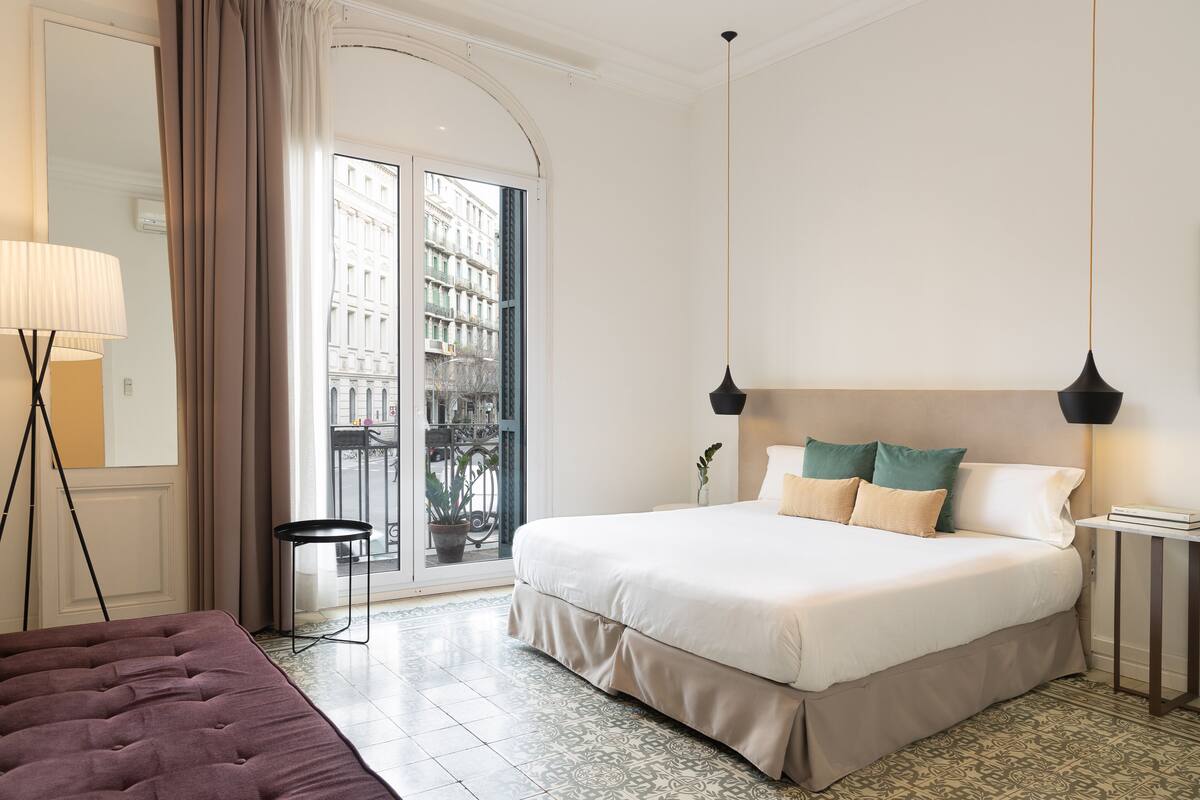 A hotel room with a large bed, a small table, a floor lamp, and a view of the city through a glass door.