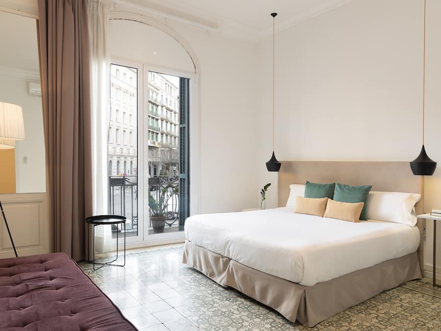 A hotel room with a large bed, a small table, a floor lamp, and a view of the city through a glass door.