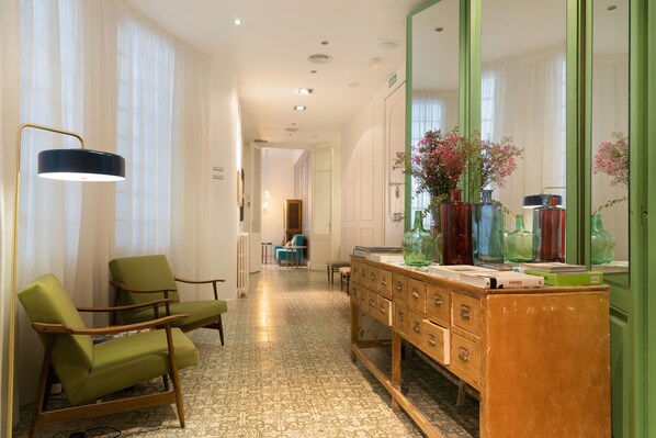 A hotel room with a green armchair, a wooden dresser with drawers, a large mirror, and a floor with patterned tiles.