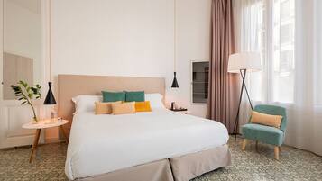Quarto de hotel com uma cama grande, uma mesinha de cabeceira, uma luminária, uma cadeira e uma janela com cortinas.