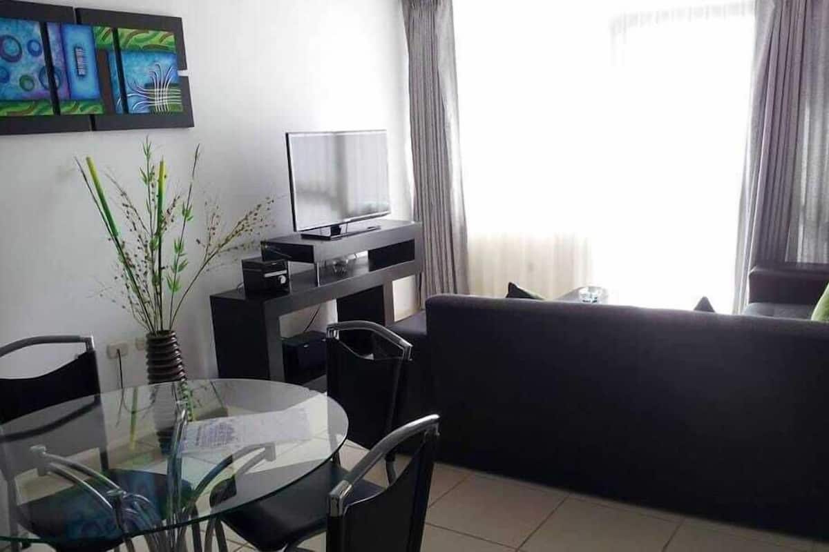 A modern living room with a glass dining table, black chairs, a black sofa, a flat-screen TV on a black stand, and abstract art on the wall.
