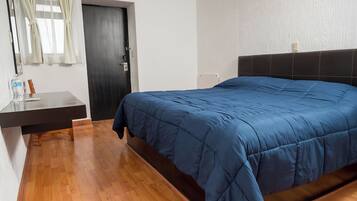 A bedroom with a wooden floor, a bed with a blue bedspread, a nightstand, a mirror, a door, and a wall-mounted light fixture.