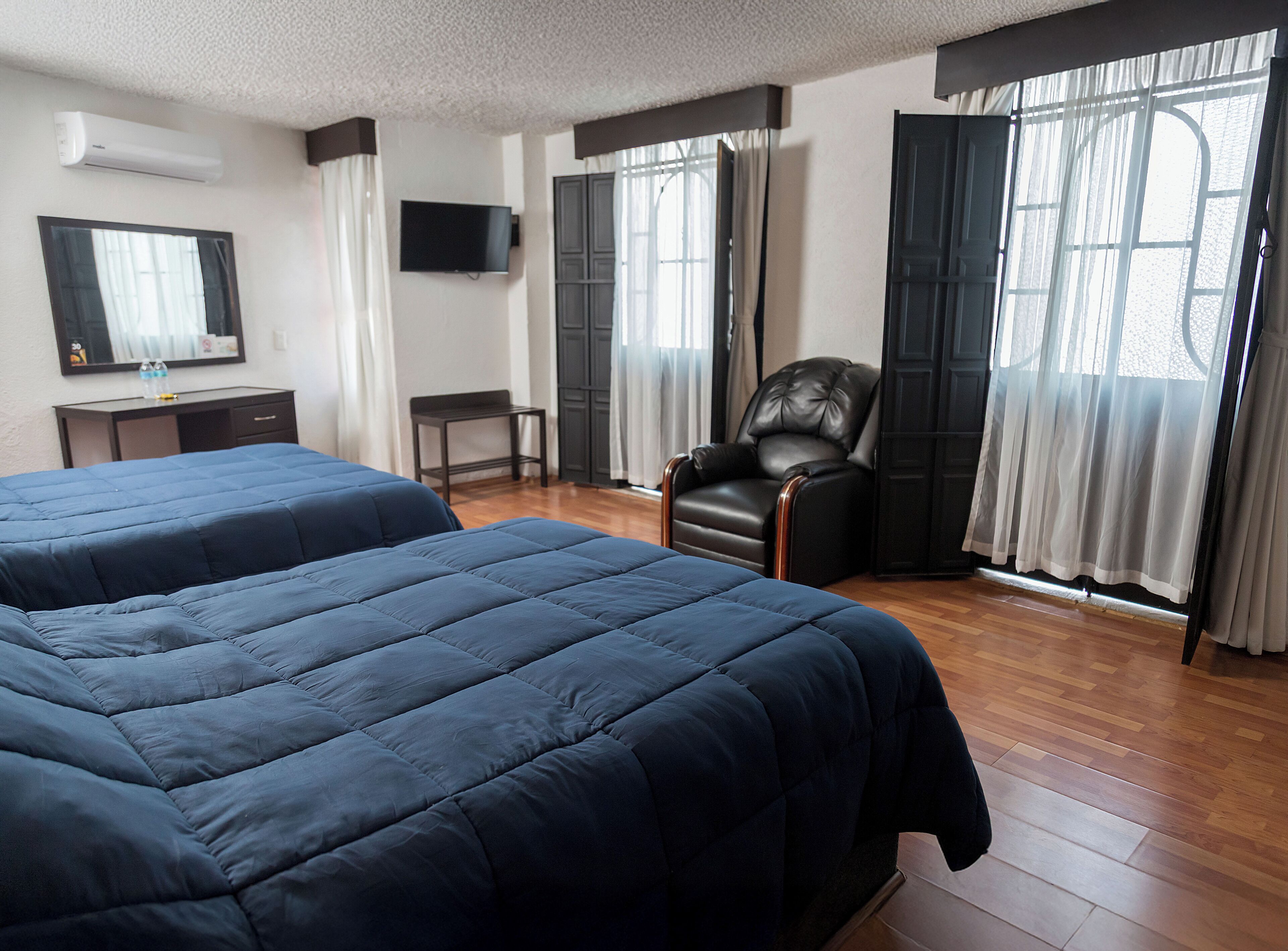 Habitación de hotel con dos camas, un escritorio, una silla, un televisor y ventanas amplias con cortinas.
