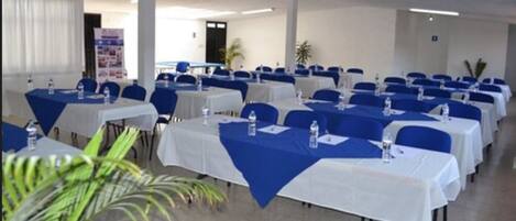 Un salón de conferencias con filas de mesas, cada una con mantel blanco y cubre-sillas azules.