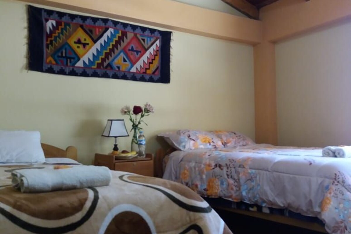 A room with two beds, wooden ceiling beams, a wall tapestry, a nightstand with a lamp, and a vase with flowers.