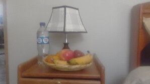 A small wooden nightstand with a lamp, a bottle of water, and a basket of fruit.