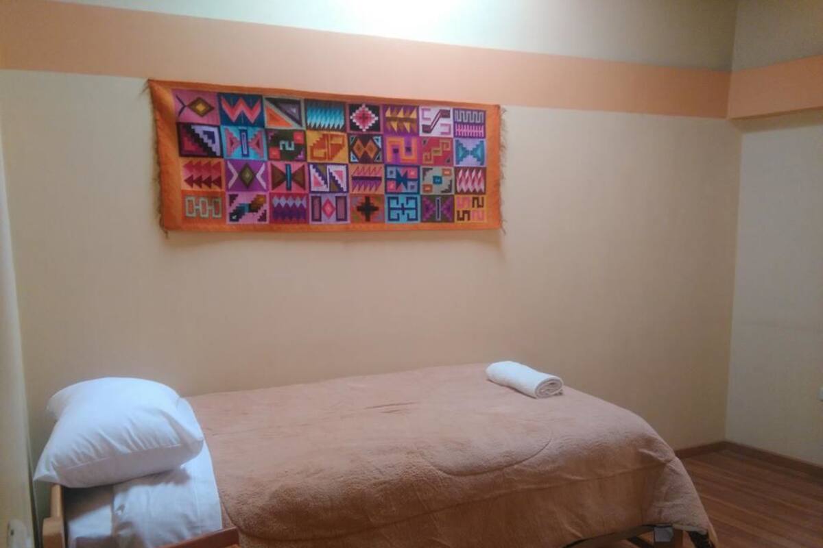 A single bed with a beige blanket and a white pillow, a wooden nightstand, and a colorful quilt hanging on the wall.