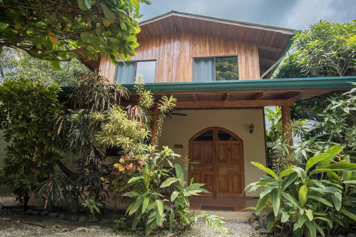 A wooden house with a green roof, surrounded by various plants and trees.