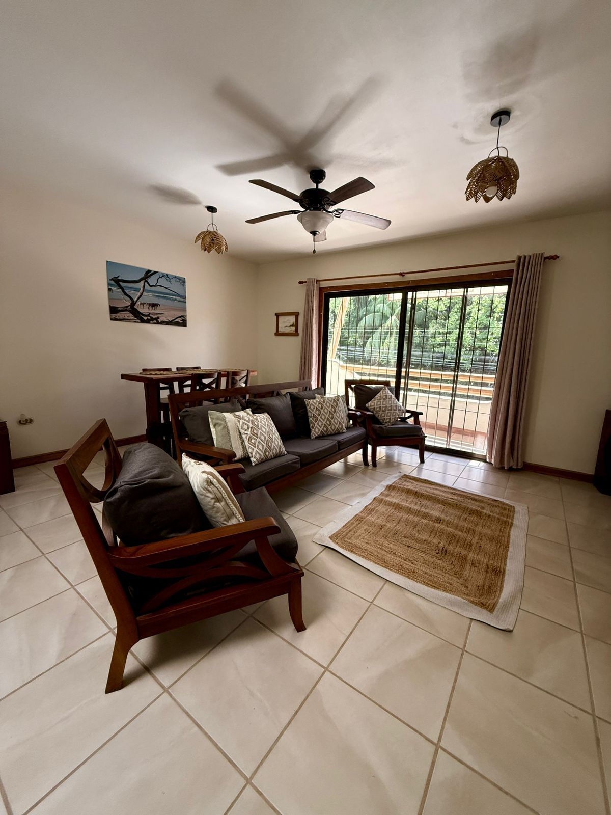 A spacious living room with a ceiling fan, a large window with curtains, a sofa set, and a dining table.