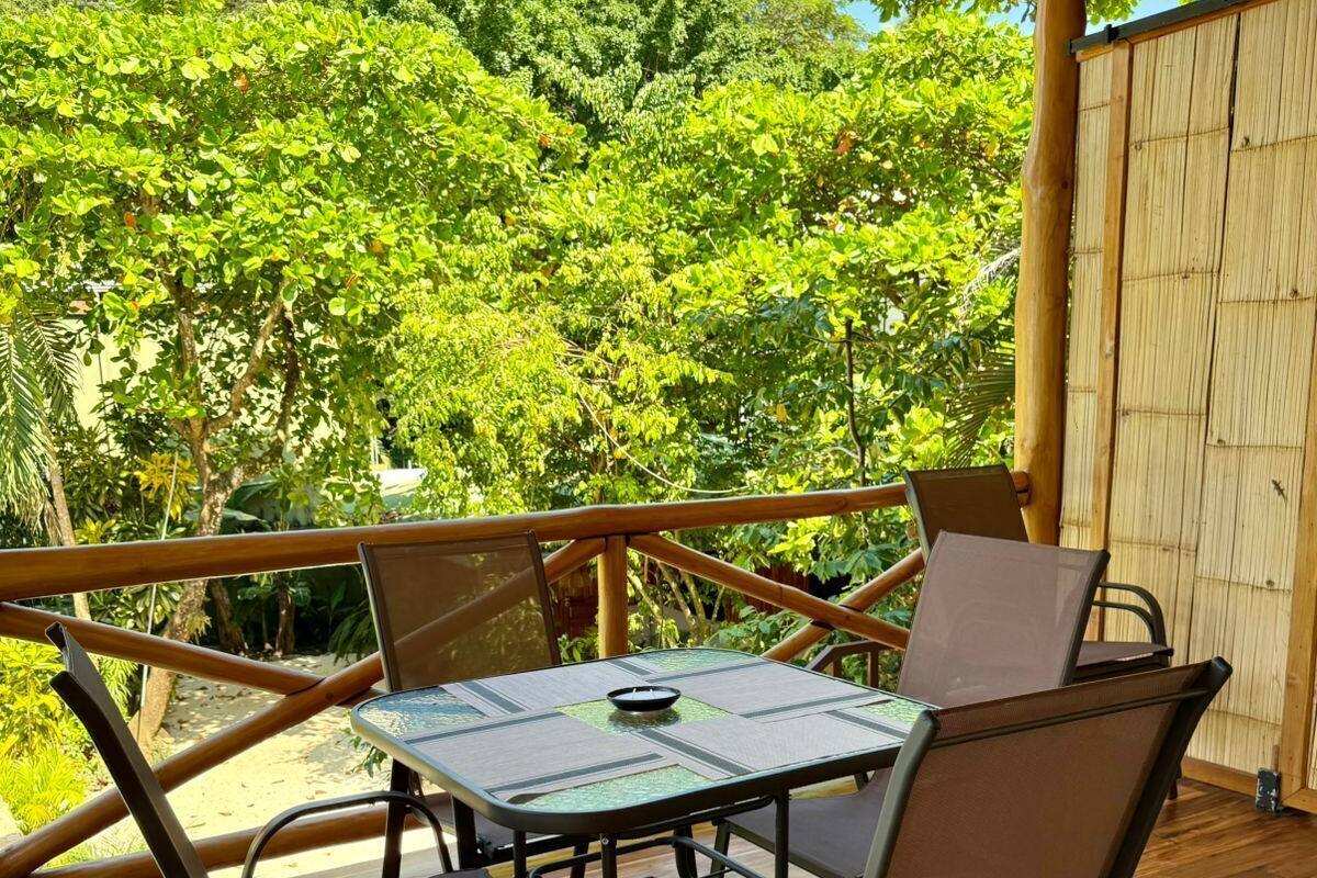 A wooden deck with a glass-top table and four chairs, surrounded by a bamboo fence, overlooking a lush garden.