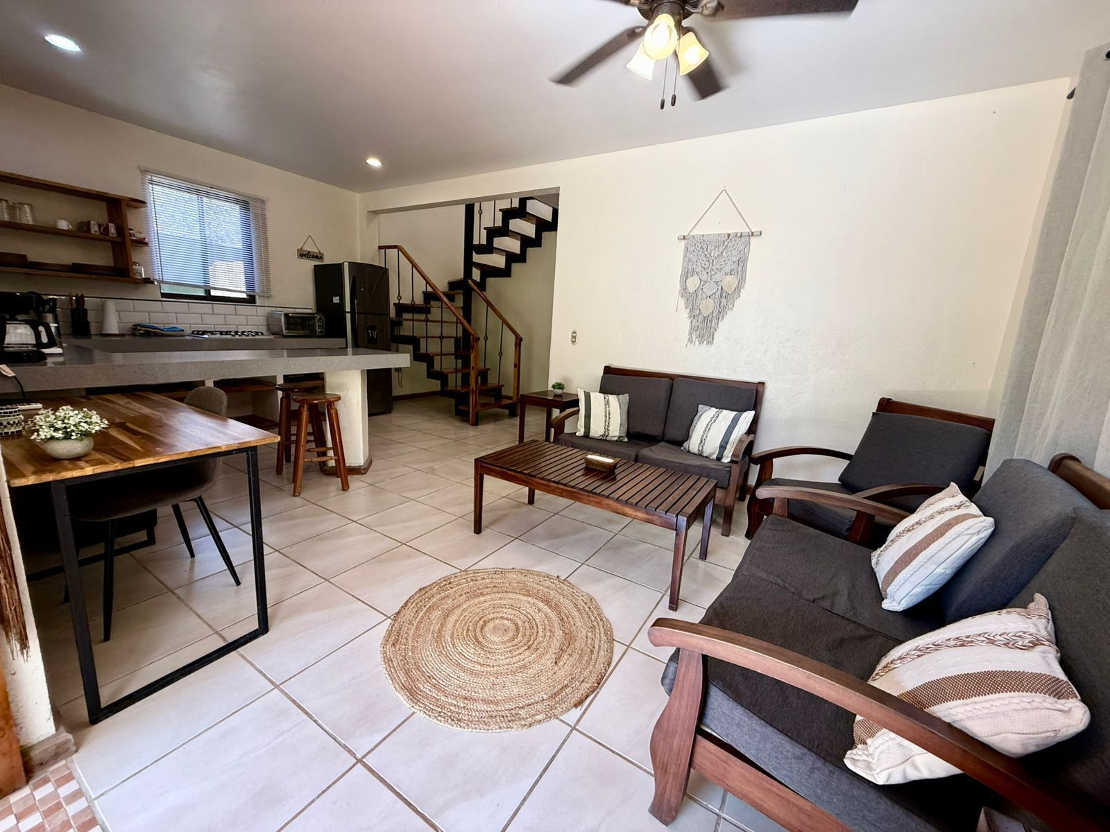 A modern living room with a sofa, coffee table, and dining area. A staircase leads to a kitchen with a window.