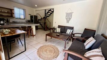 A modern living room with a sofa, coffee table, and dining area. A staircase leads to a kitchen with a window.