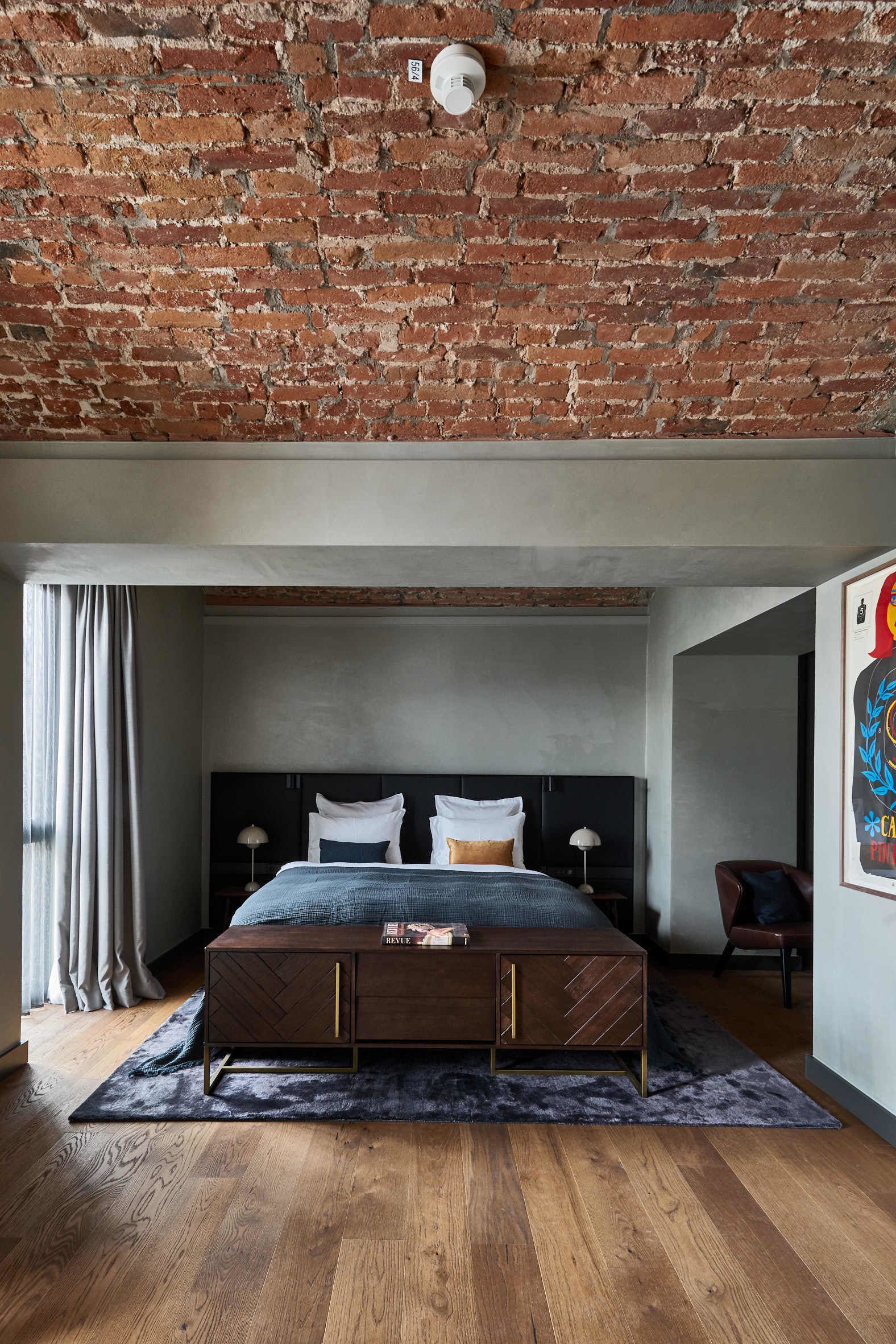Een slaapkamer met een bakstenen plafond, een bed met een houten hoofdeinde, een donkere nachtkastje en een lijst met een afbeelding aan de muur.