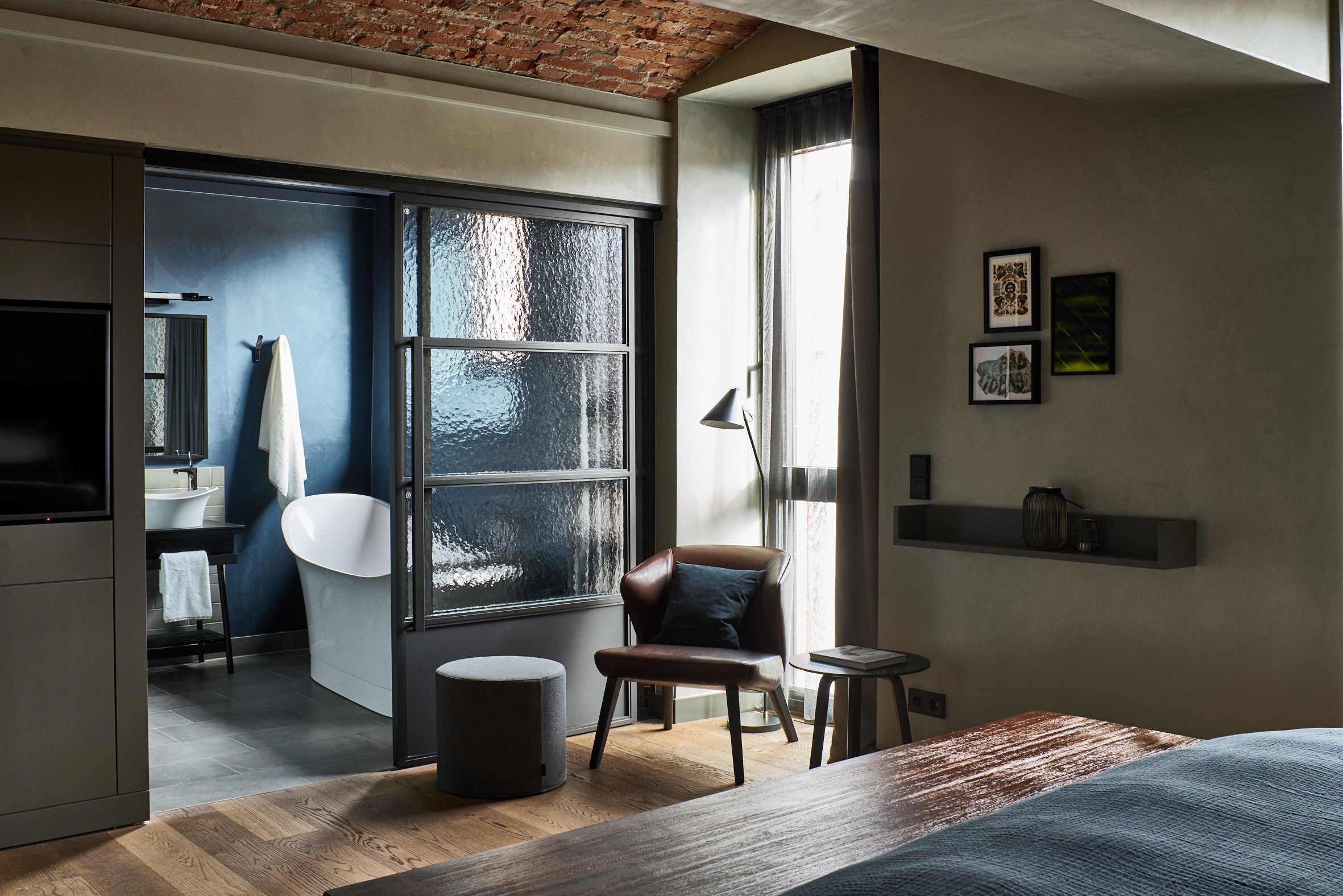Une chambre d’hôtel moderne avec une grande fenêtre, un plancher en bois, un lit, une chaise, une petite table et une salle de bain visible à travers une porte ouverte.