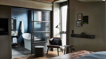 Une chambre d’hôtel moderne avec une grande fenêtre, un plancher en bois, un lit, une chaise, une petite table et une salle de bain visible à travers une porte ouverte.