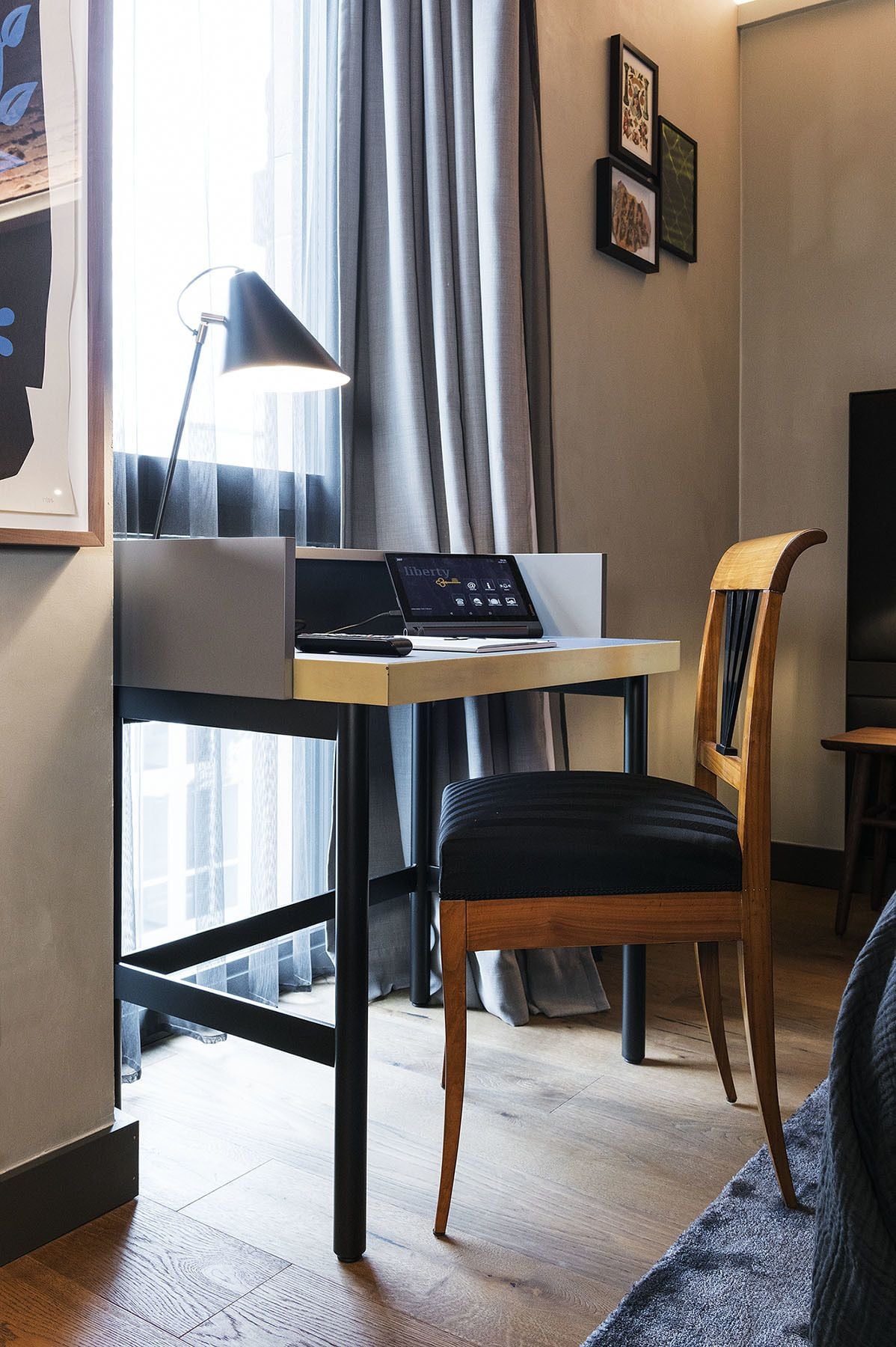 A modern room with a desk, laptop, and a chair. There is a window with curtains, a lamp, and framed pictures on the wall.