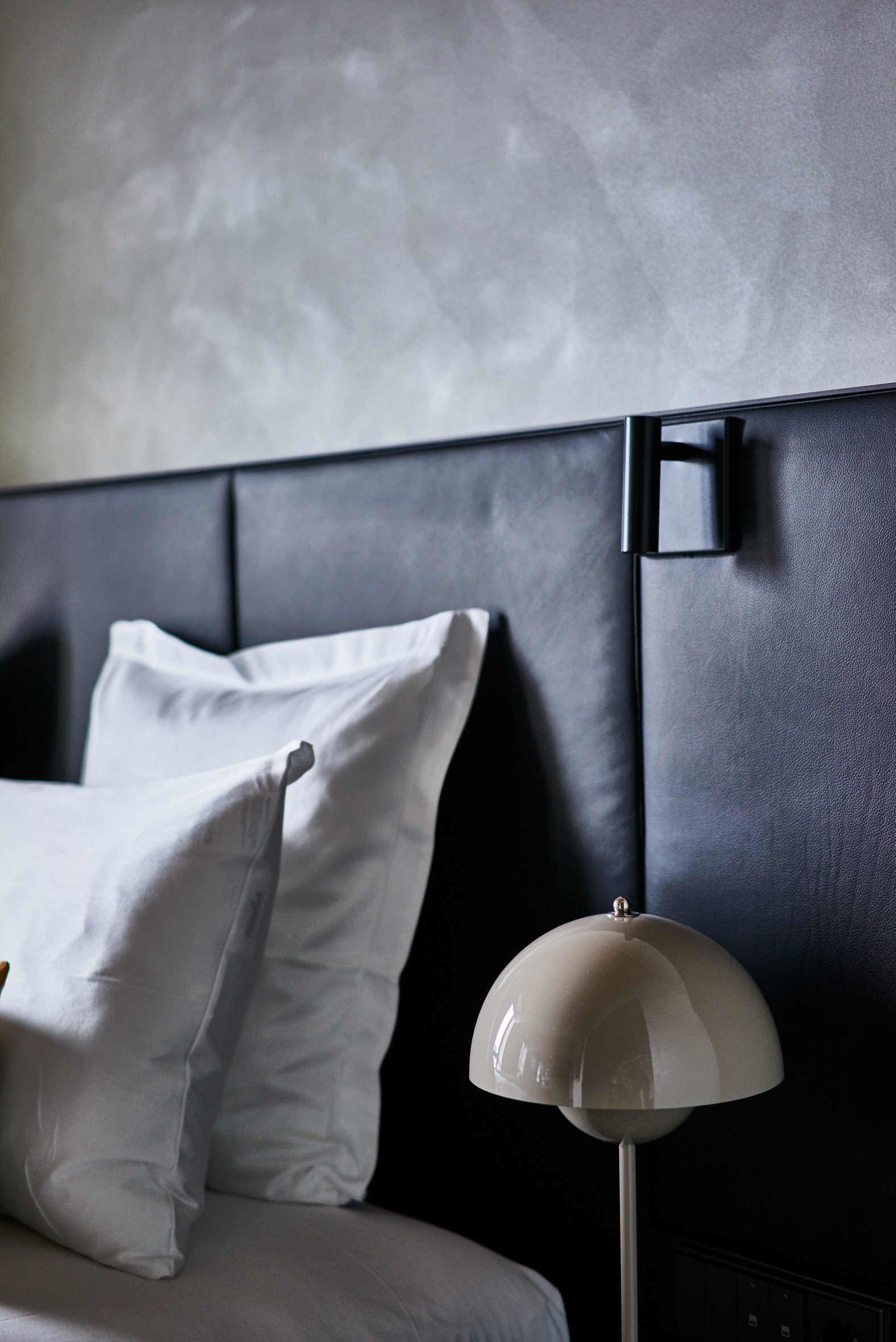 A modern hotel room with a bed, white pillows, a black headboard, and a white floor lamp.