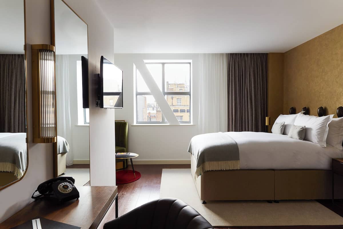 Une chambre d’hôtel avec un grand lit, un bureau avec un téléphone, une chaise, une petite table et une fenêtre avec des rideaux.
