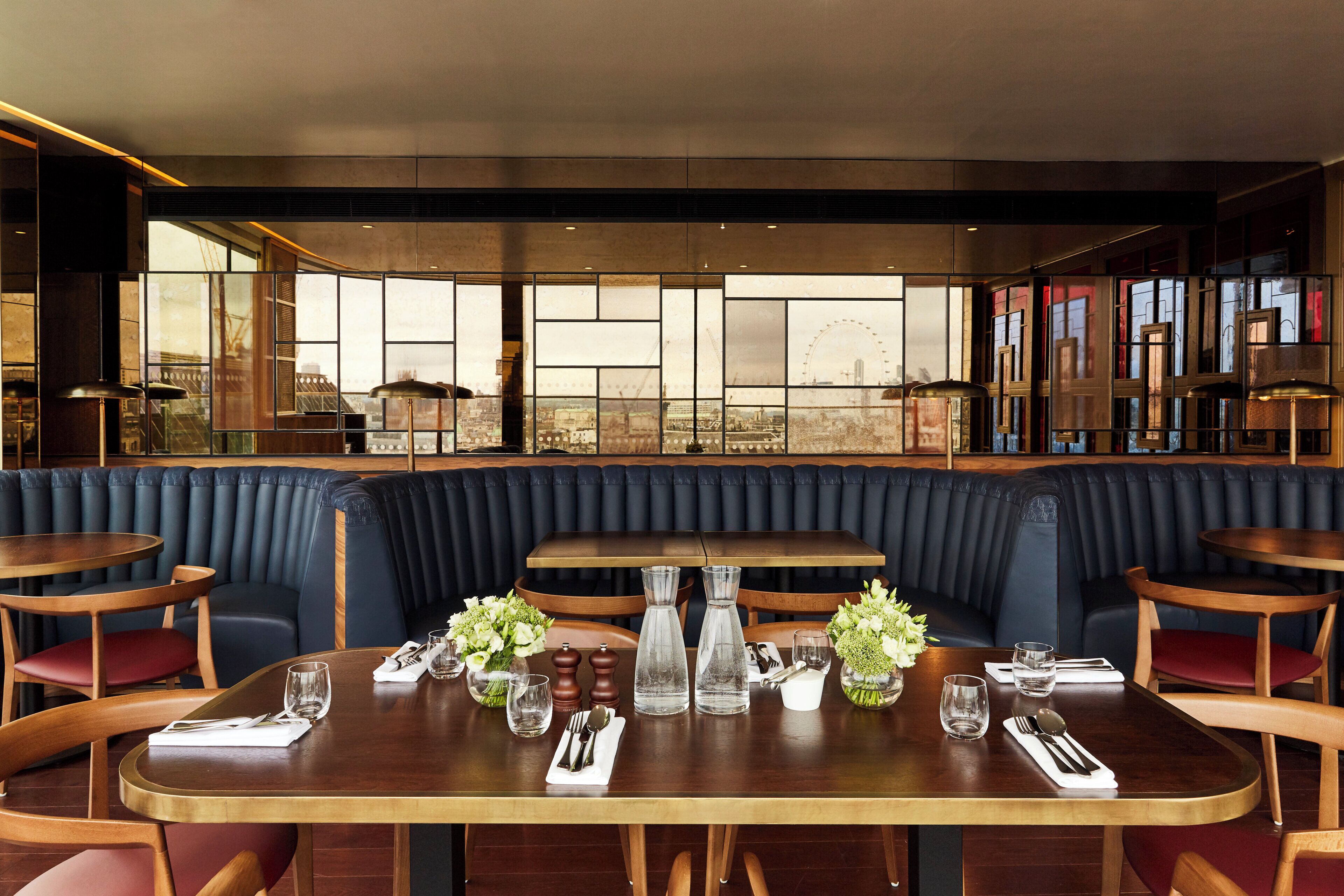 A restaurant with a long table set for dining, cushioned seating, and a view of a cityscape through large windows.