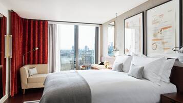 Habitación de hotel moderna con una cama grande, un sofá, un escritorio y vistas de la ciudad a través de amplias ventanas.