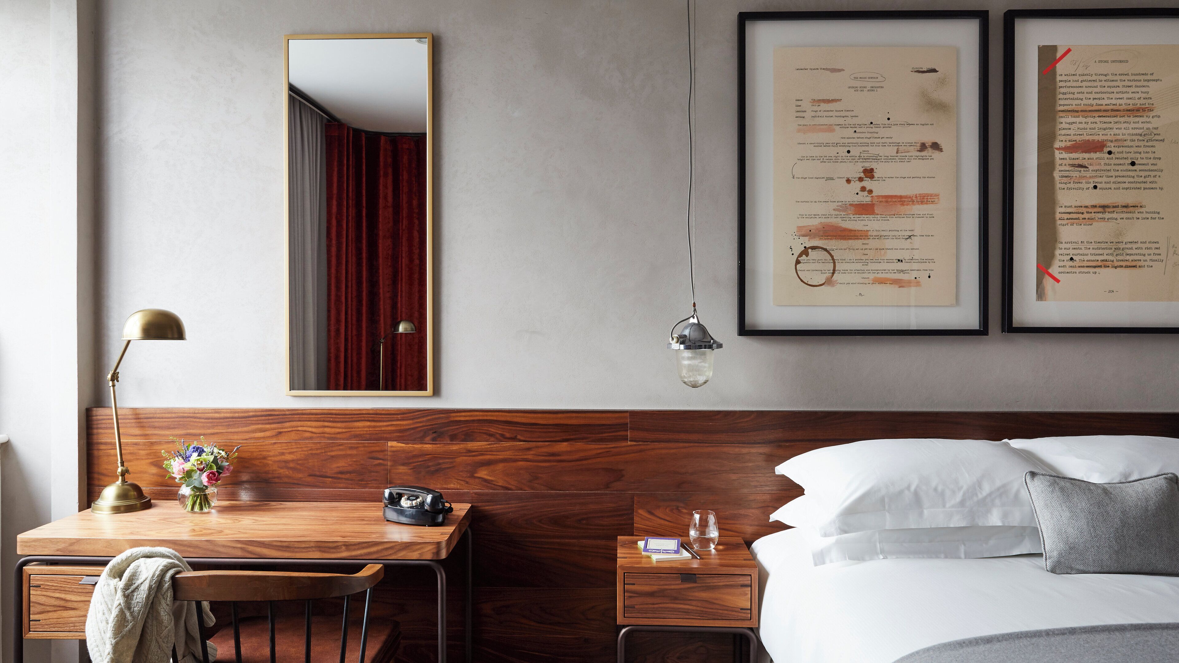A hotel room with a wooden headboard, a desk with a telephone and a vase of flowers, a mirror, and two framed documents on the wall.
