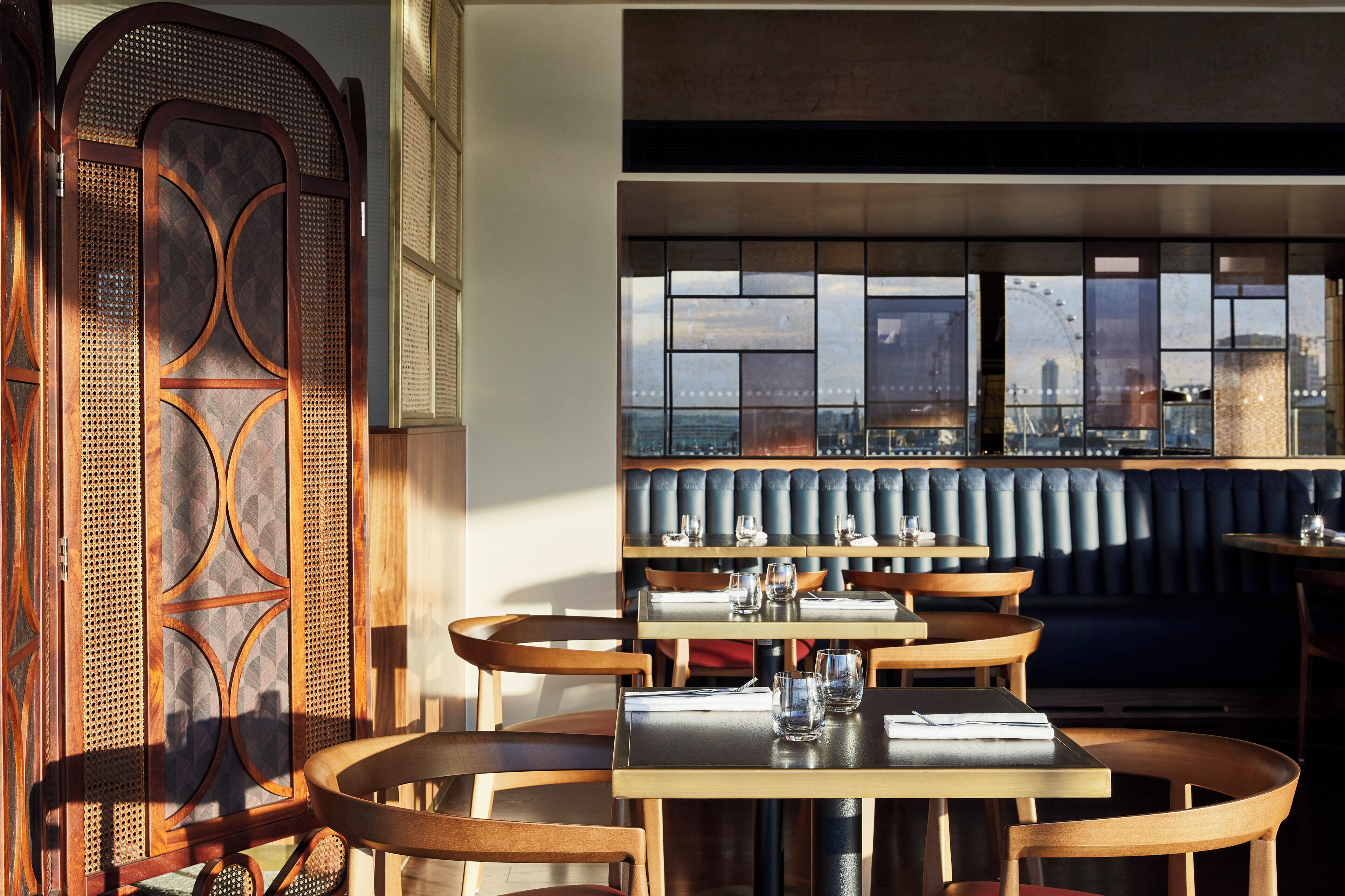 A restaurant interior with wooden furniture, a large window with a city view, and a decorative wooden panel on the left.