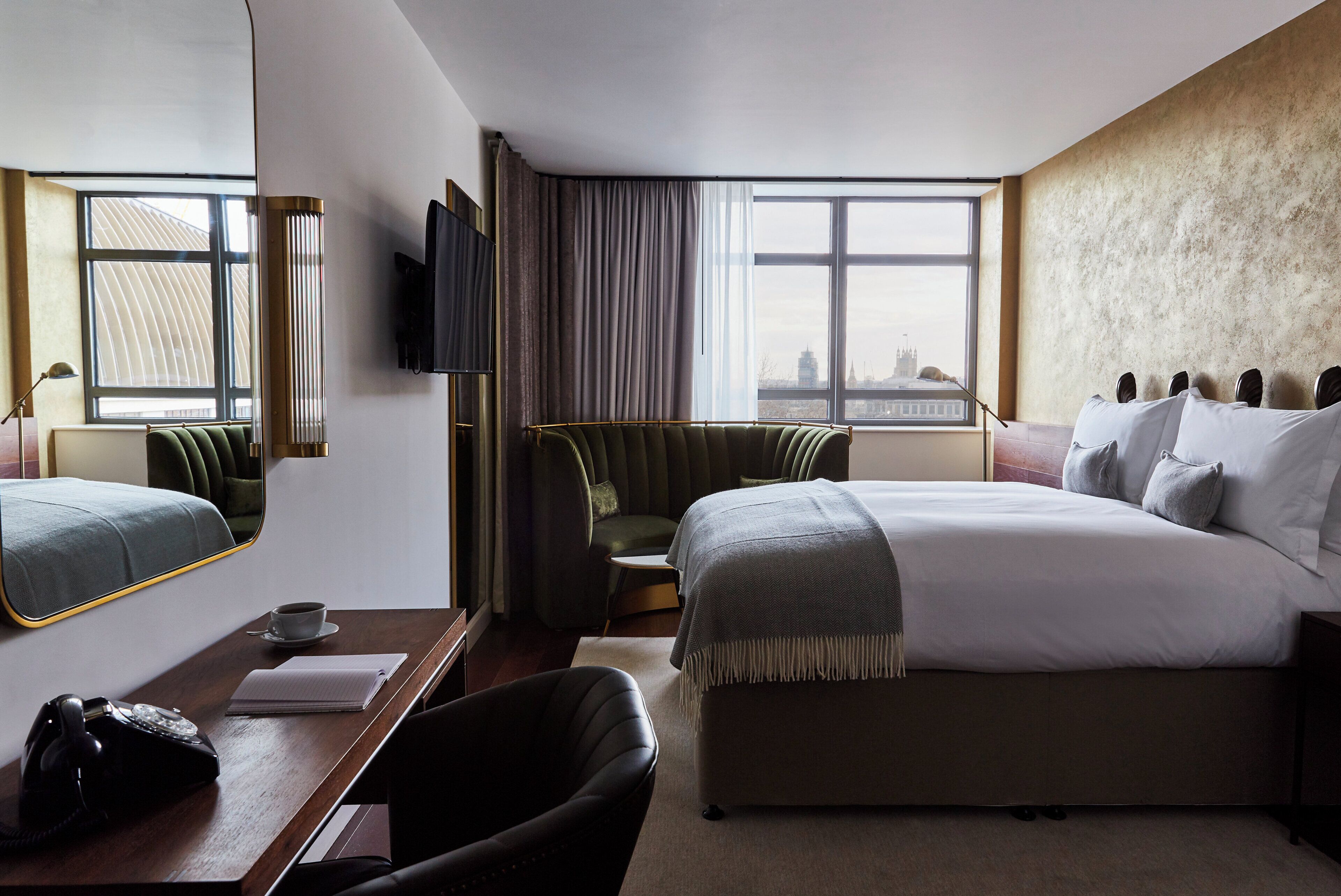 A hotel room with a bed, a desk with a telephone, a chair, a television, and a view of the city through large windows.