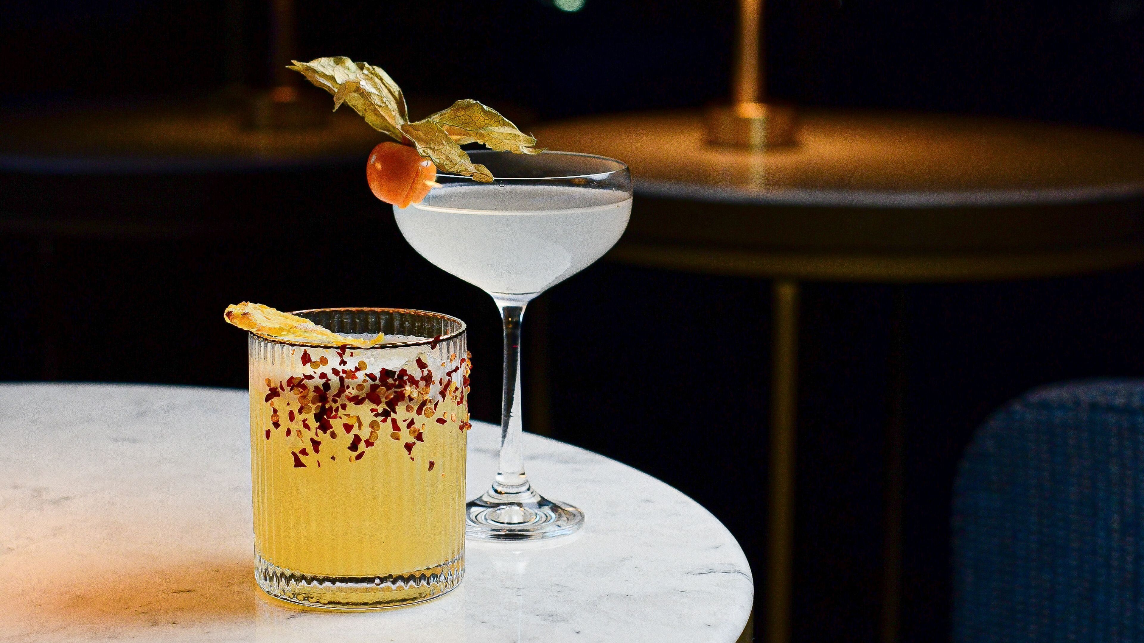 A cocktail in a martini glass garnished with a leaf and a slice of orange, and a highball glass with a slice of orange and red pepper flakes.