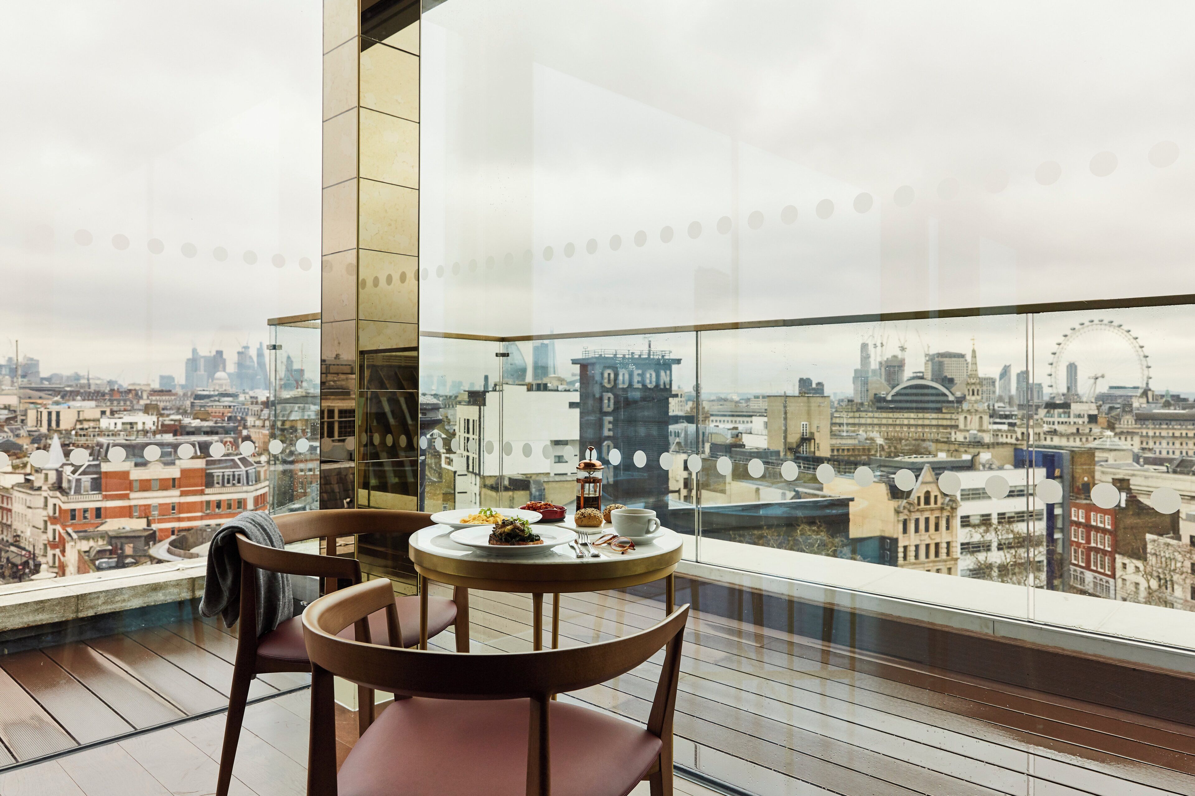 A dining area with a table and chairs, a city view through a large window, and a building with 'ODEON' written on it.