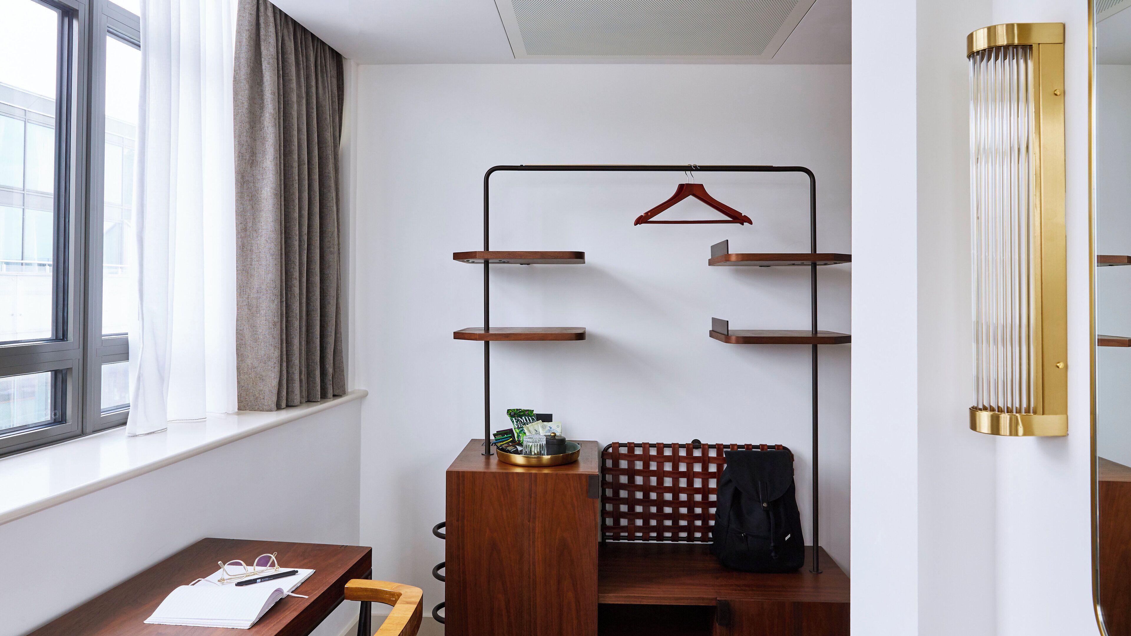 A compact hotel room with a wooden desk, a hanging coat rack, and a radiator.