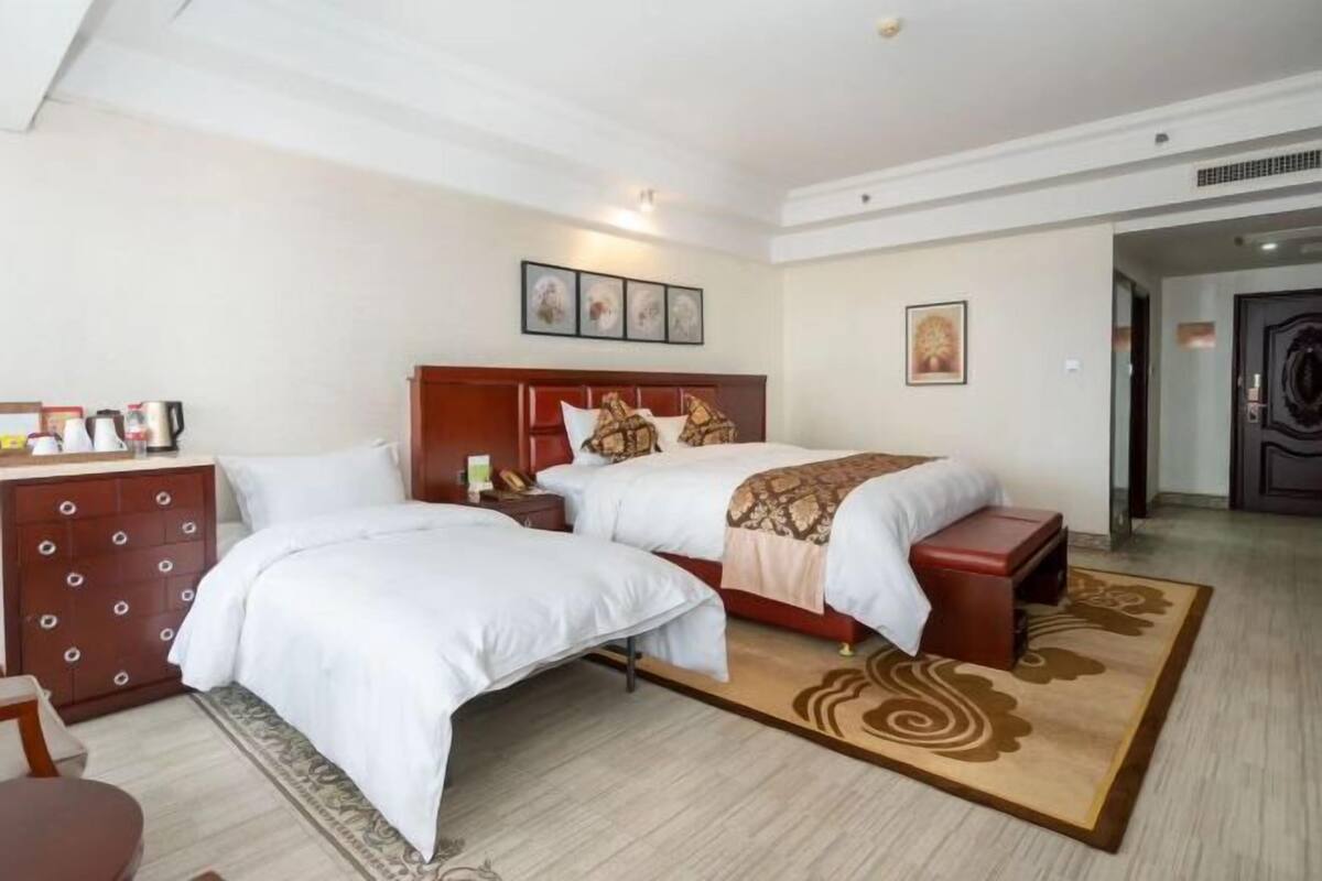 A hotel room with two beds, a wooden headboard, a desk with a chair, and a small table with a lamp.