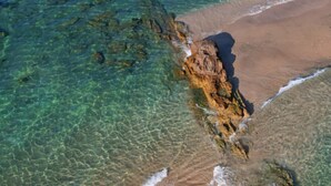 Vista aerea di una spiaggia con acque limpide, un promontorio roccioso e una riva sabbiosa.