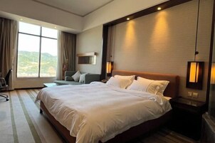 Quarto de hotel com cama grande, escrivaninha e cadeira. Há uma janela com cortinas, luminária na parede e vista para o exterior.