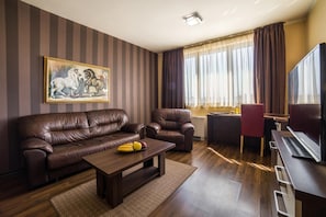 A living room with a brown leather sofa, a coffee table with fruits, a painting, and a large flat-screen TV. - Business Hotel Plovdiv (Plovdiv)