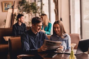 A person reading a newspaper at a table in a cafe with another person seated beside them.
