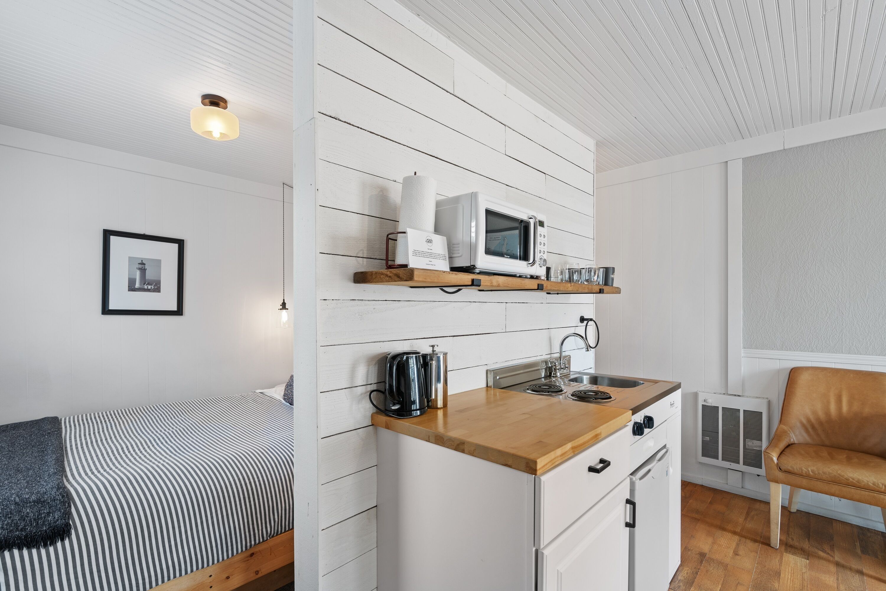 A compact kitchen with a microwave, kettle, and sink, adjacent to a bed with a striped blanket.