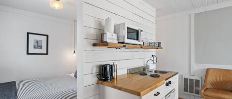 A compact kitchen with a microwave, kettle, and sink, adjacent to a bed with a striped blanket.