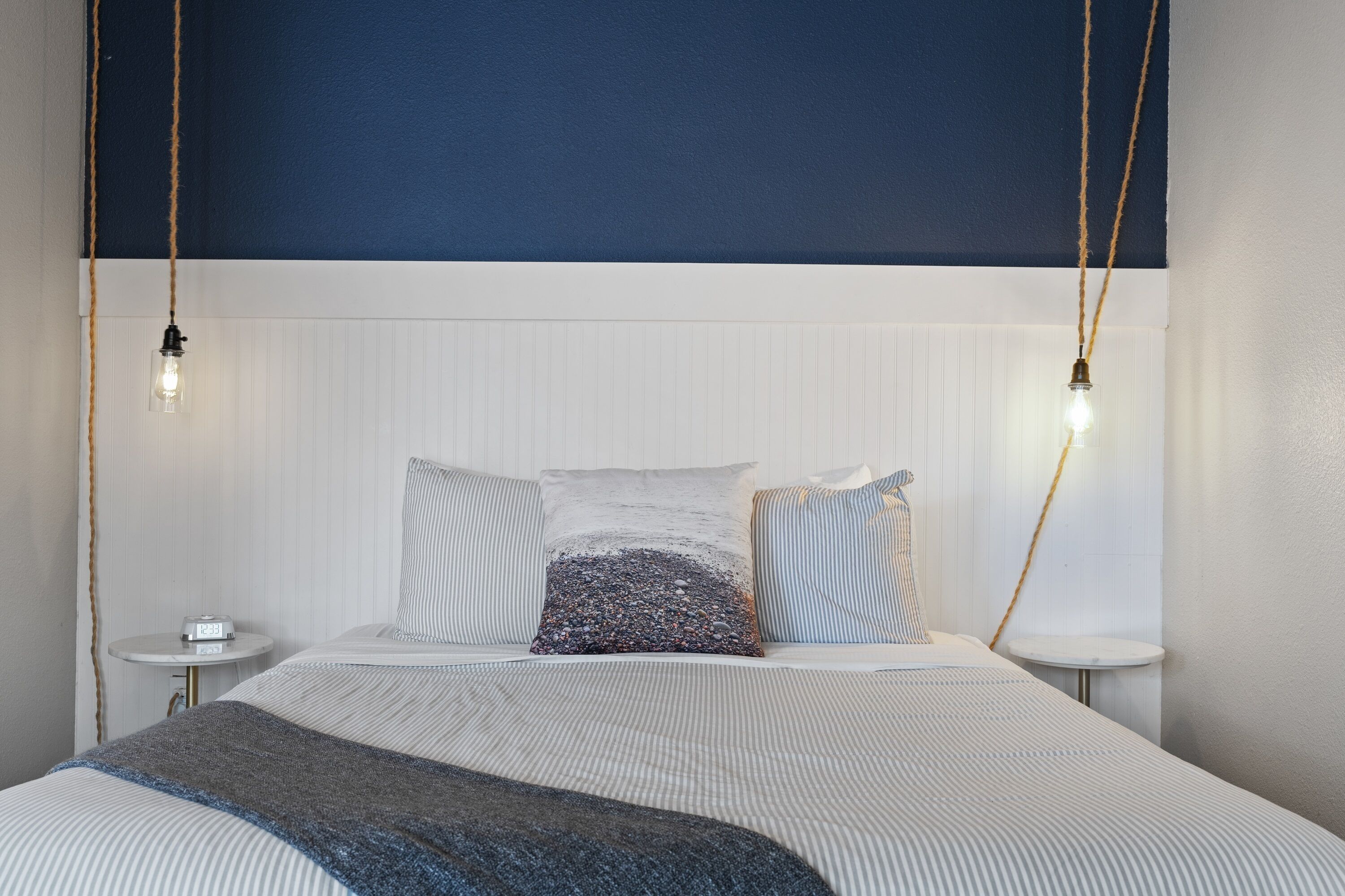 A neatly made bed with striped bedding and a textured throw blanket. There are two bedside tables with a small clock on one. The room features a white headboard and a dark blue accent wall.