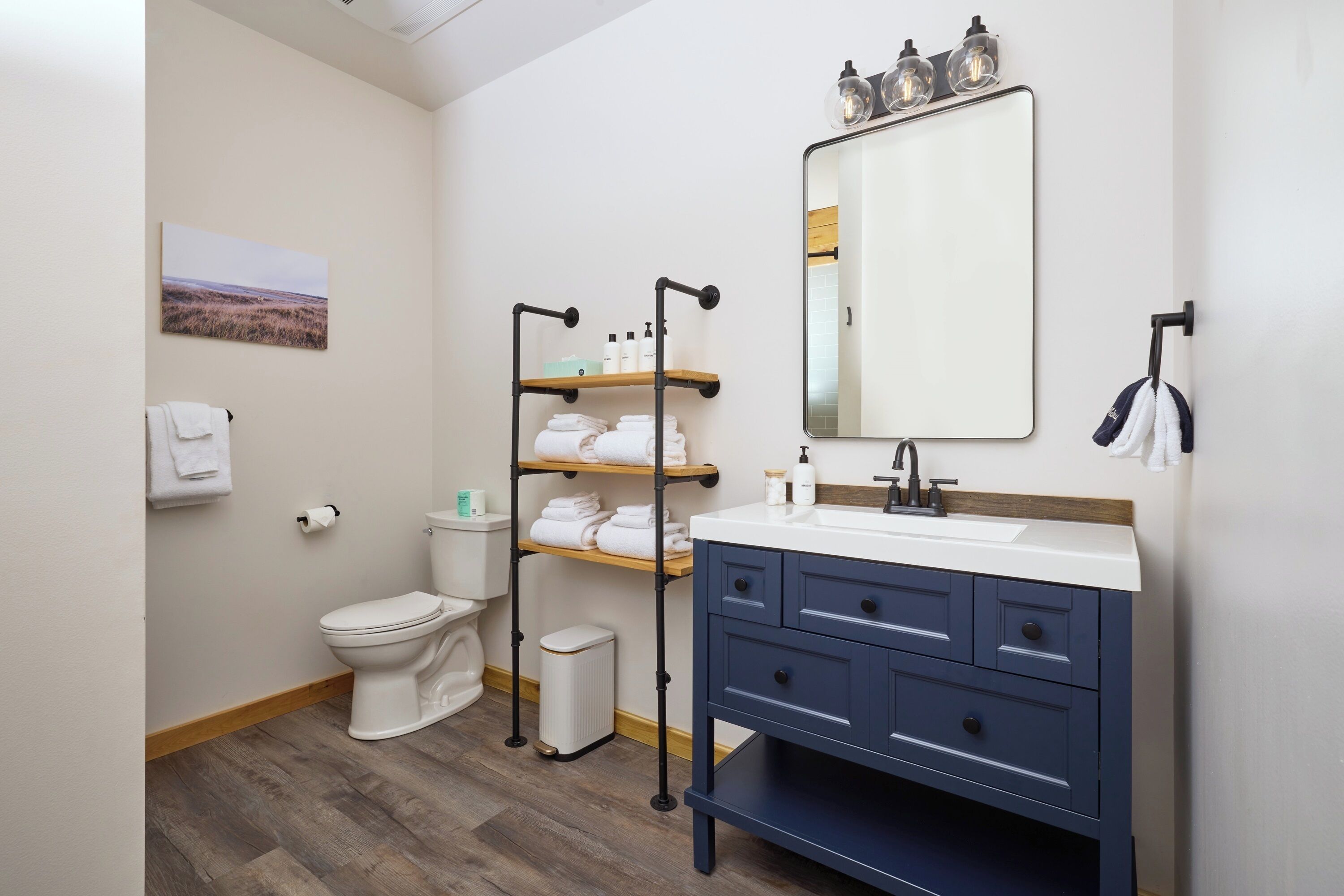 A modern bathroom with a white toilet, a blue vanity with drawers, a mirror with lights, and a towel rack.