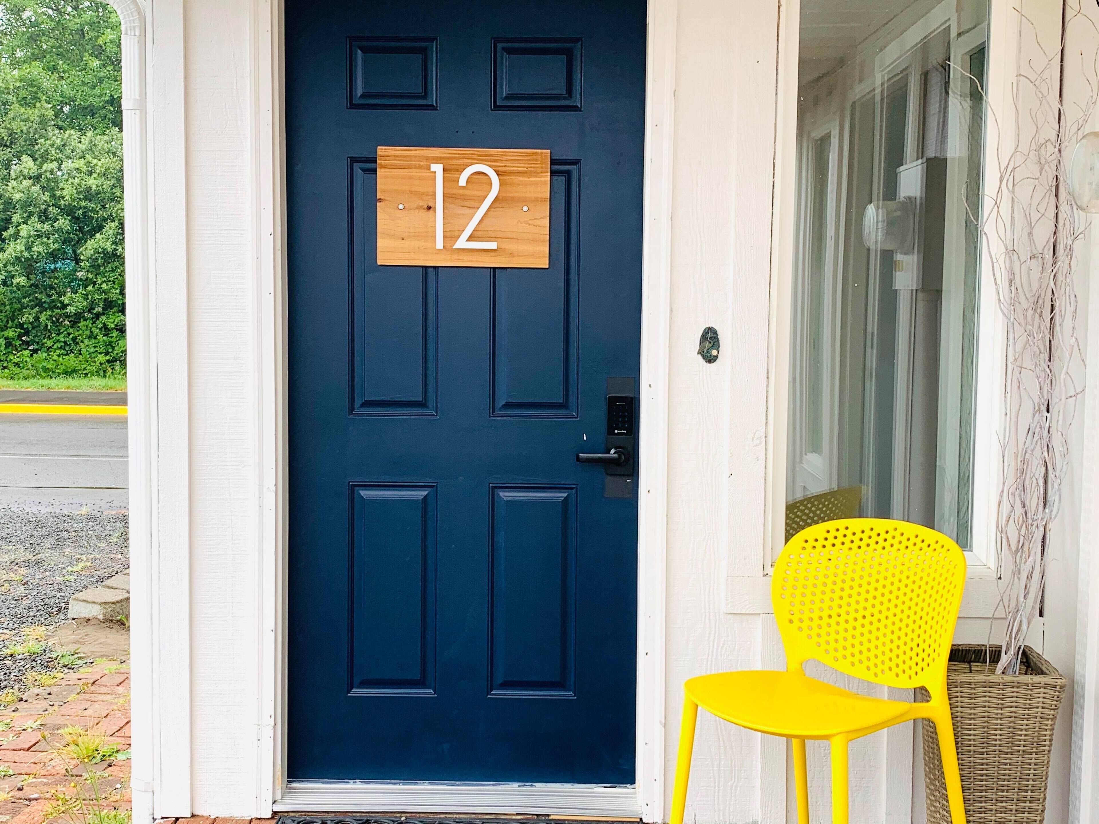 Una puerta azul oscuro con una placa de madera con el número 12 y una silla amarilla afuera.