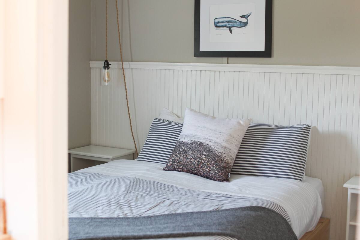A neatly made bed with striped bedding and a textured pillow, a framed whale illustration on the wall, and a simple bedside table.