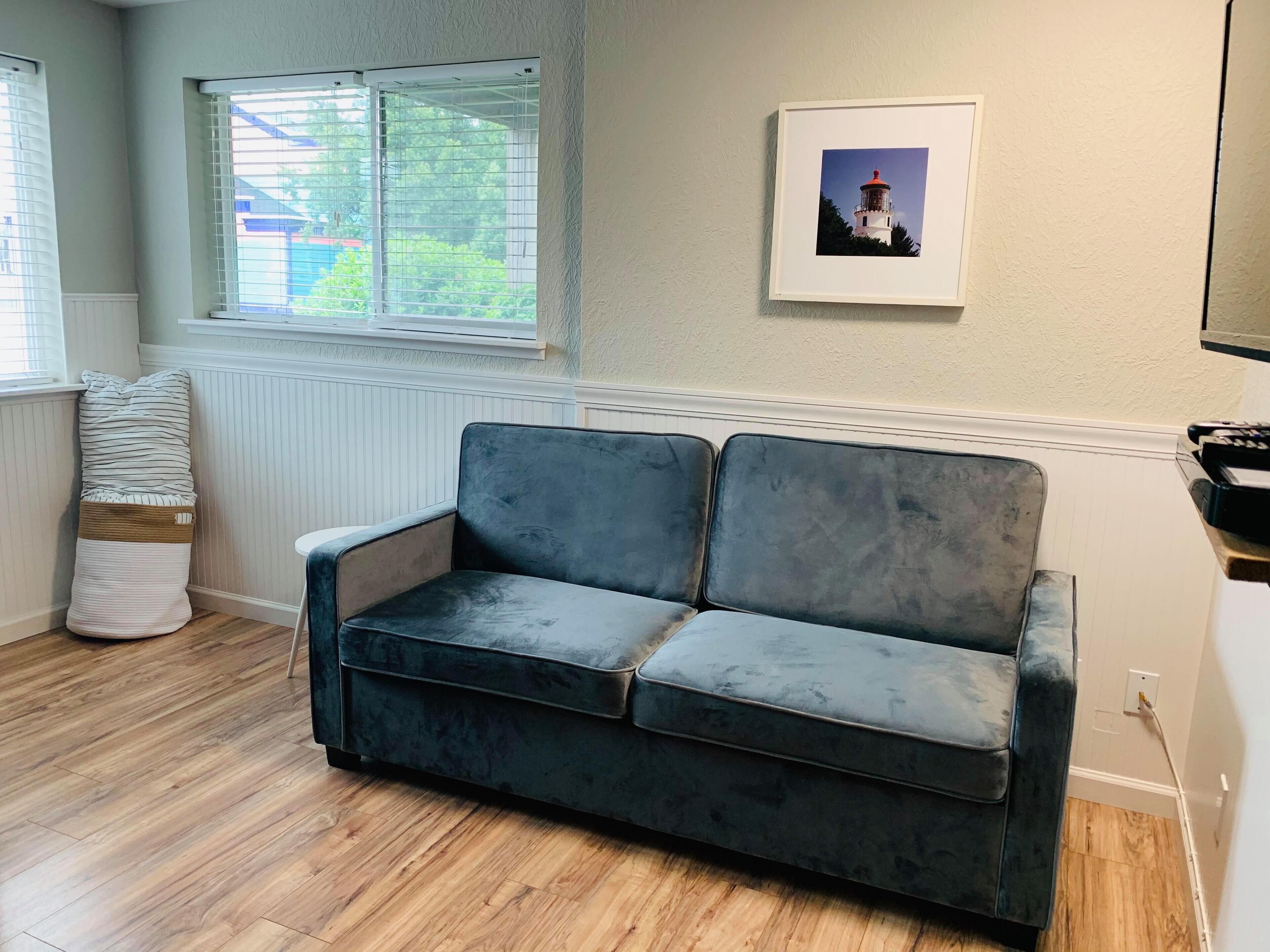 Ein Zimmer mit einem dunkelblauen Sofa, einem Fenster mit Jalousien, einer Heizung, einem gerahmten Bild an der Wand und einem Holzboden.