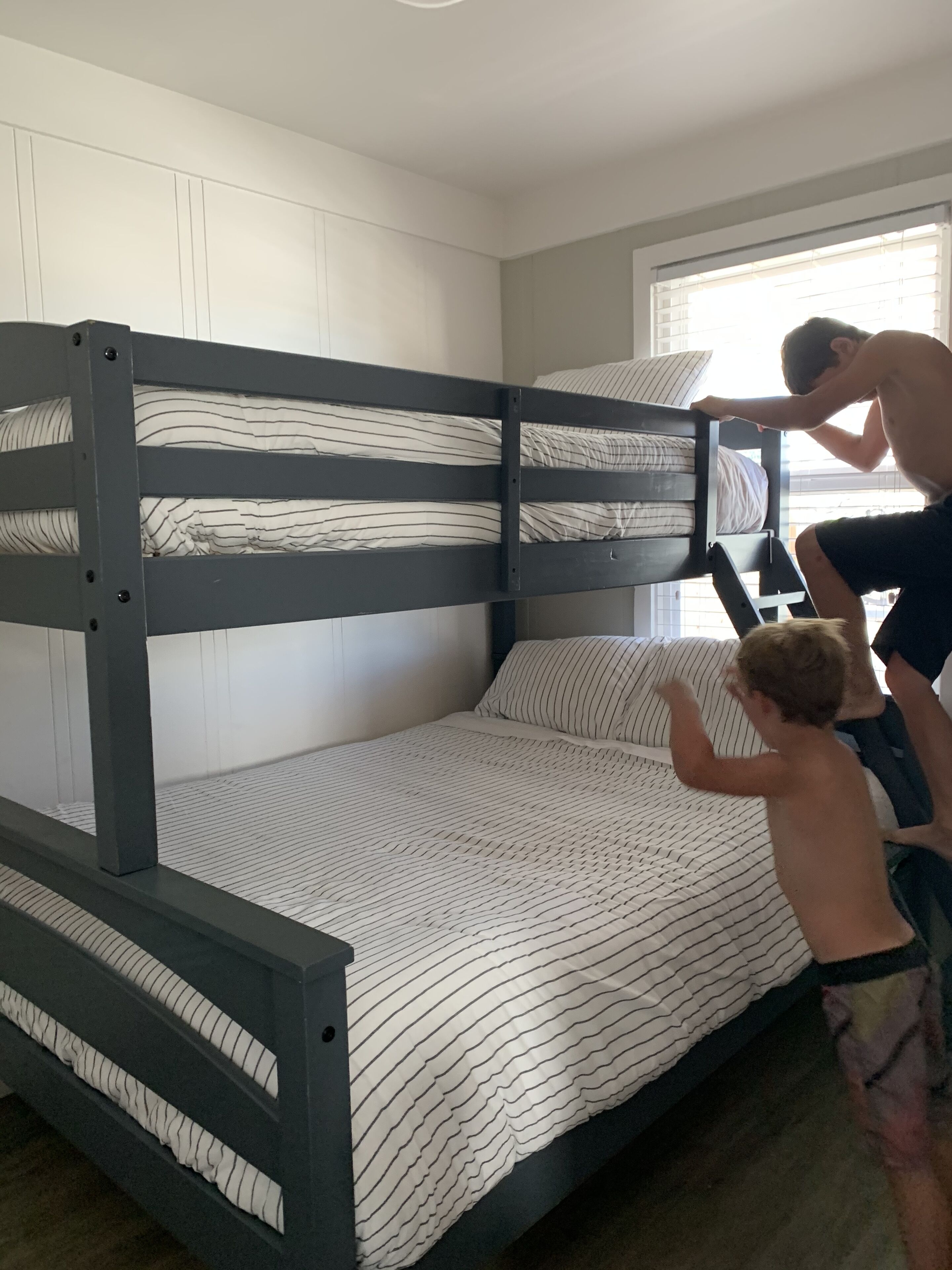 A bunk bed with a striped mattress and pillows, a wooden frame, and a window with blinds.