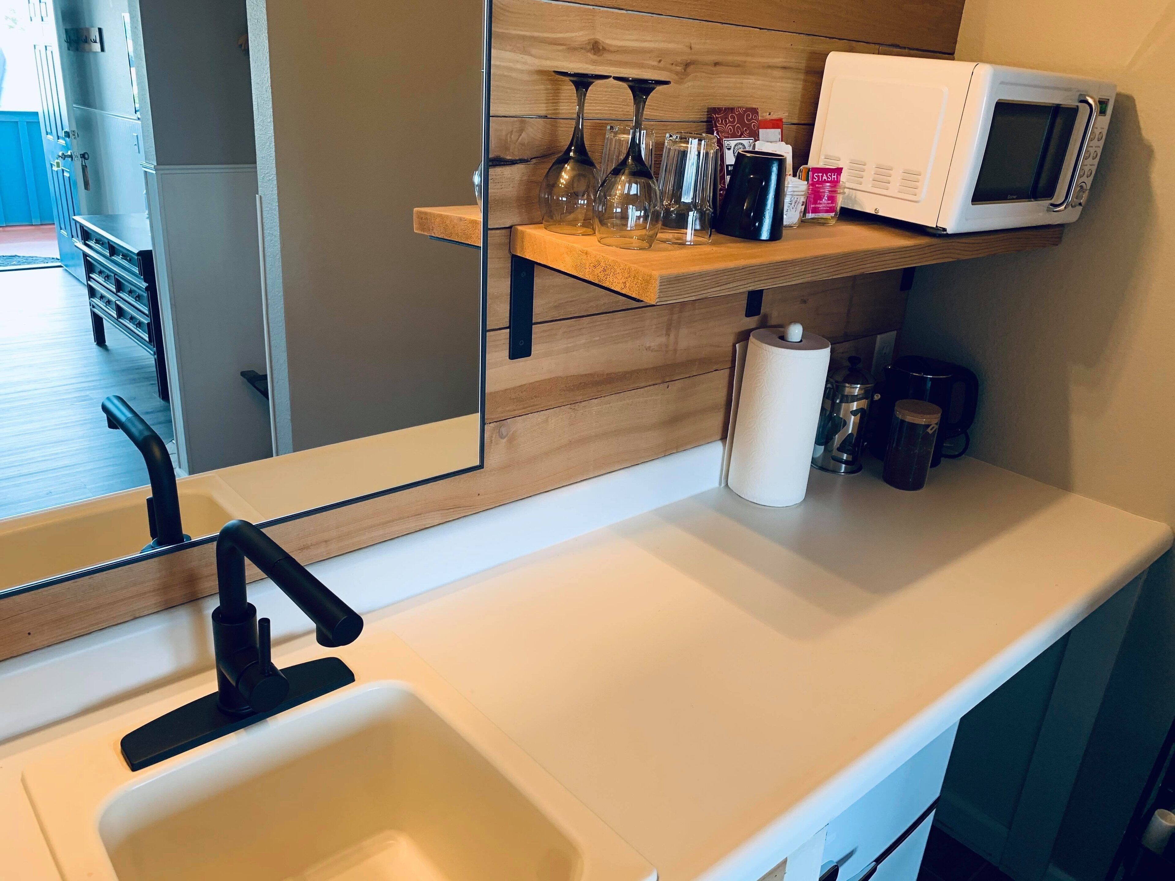 A modern kitchen with a white countertop, a black faucet, a microwave, and a wooden shelf with various items.