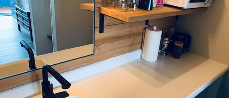 A modern kitchen with a white countertop, a black faucet, a microwave, and a wooden shelf with various items.