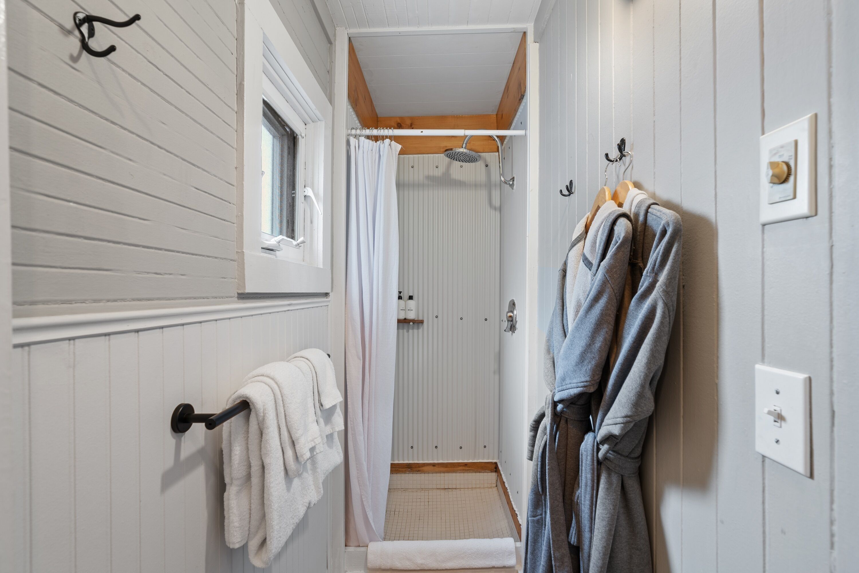 A narrow bathroom with a shower, a towel rack, and a robe hanging on the wall.