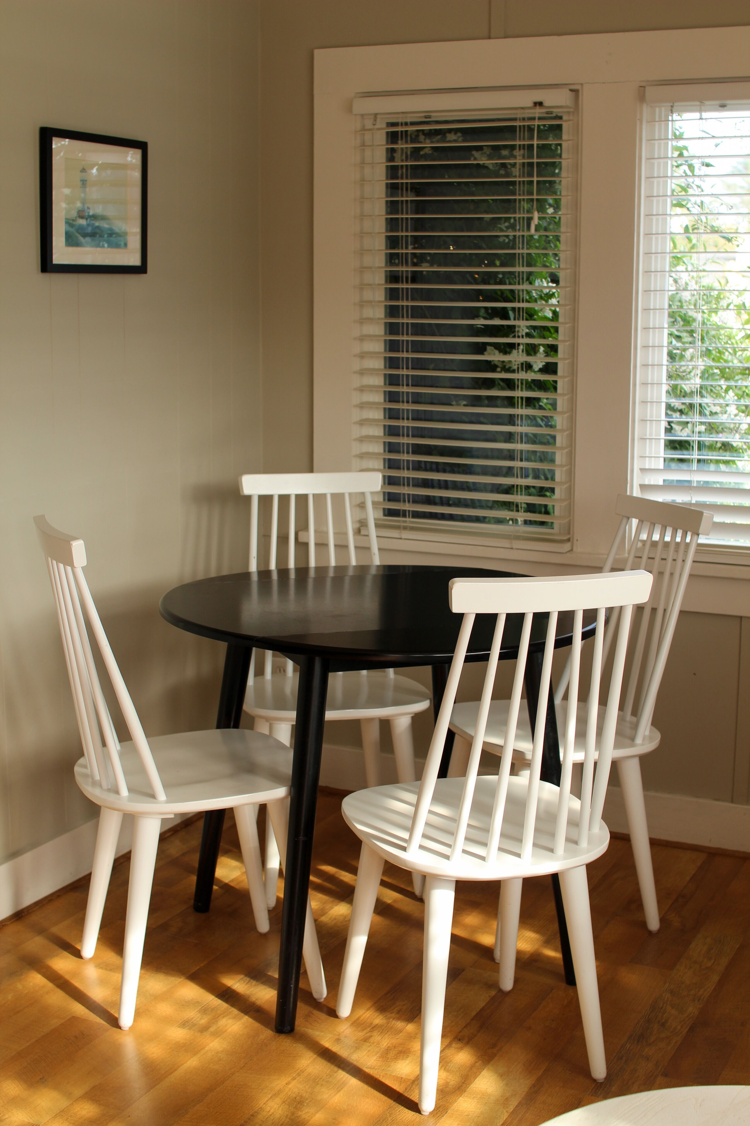 A dining area with a round table and four white chairs. There is a window with blinds, a framed picture on the wall, and a door with a window.