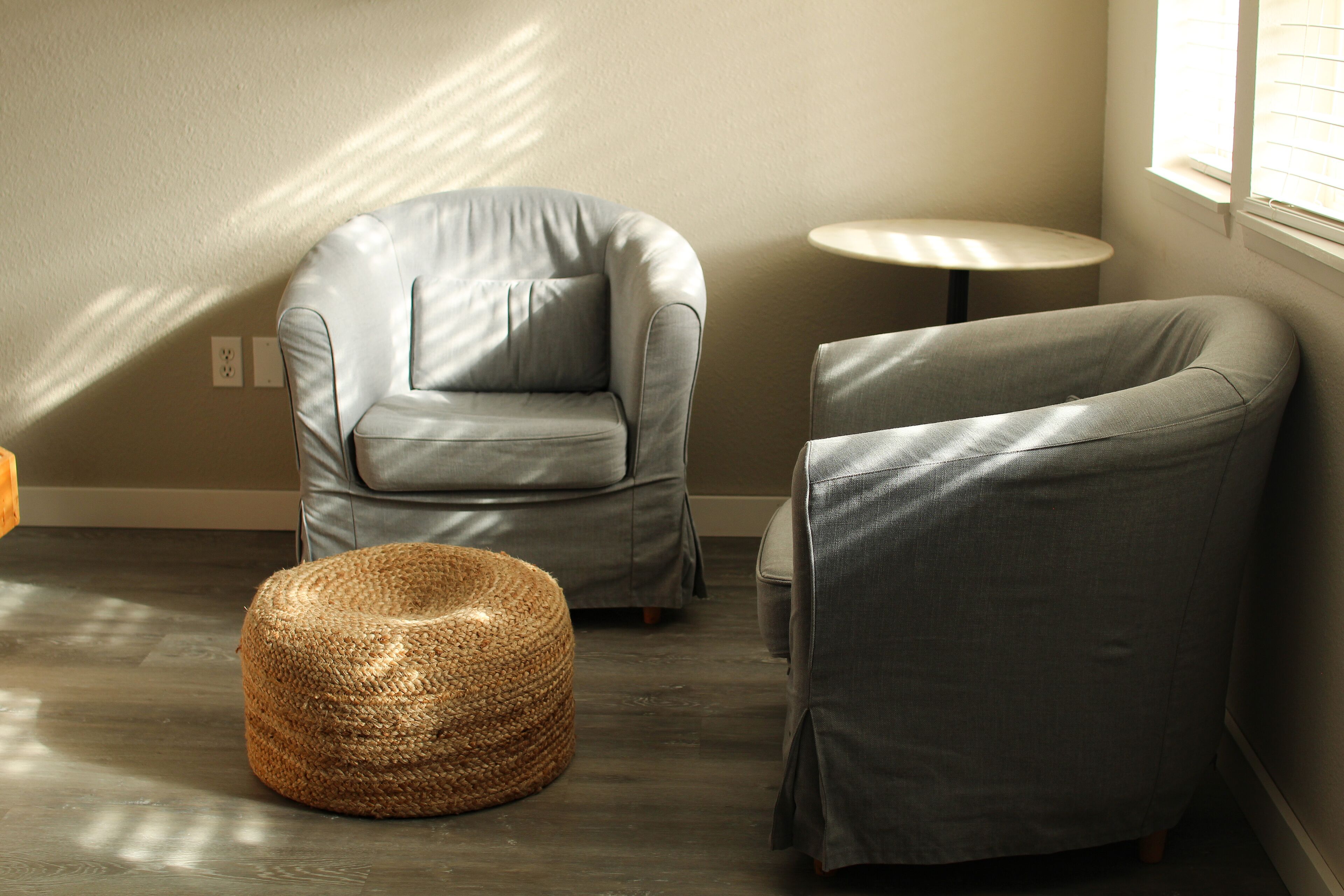 Una habitación con un sillón gris, un taburete redondo tejido, una mesita redonda y una ventana con persianas.